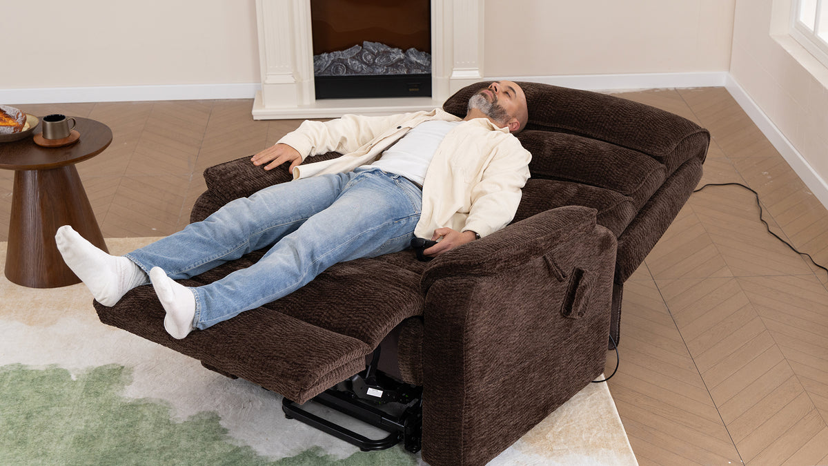 A man lying fully flat in a dark brown chenille Irene House lift recliner beside a fireplace