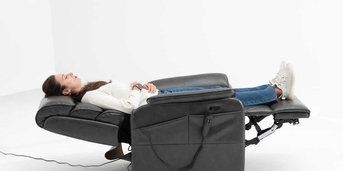 Side view of a woman lying fully flat at 180 degrees in an Irene House dark gray chenille lift recliner, head to heels supported