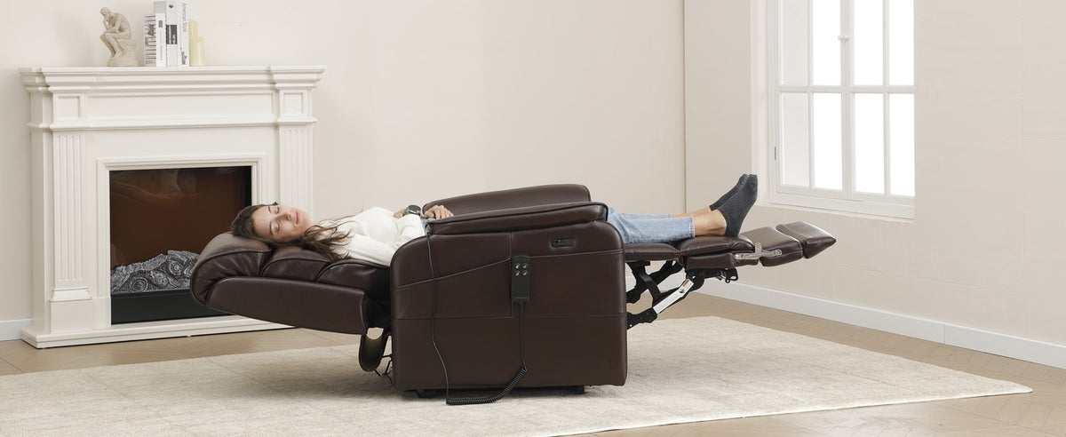 A woman lying fully flat in an Irene House 9188T tall lift recliner in dark brown leather, extended footrest supporting her legs to the heel, beside a fireplace